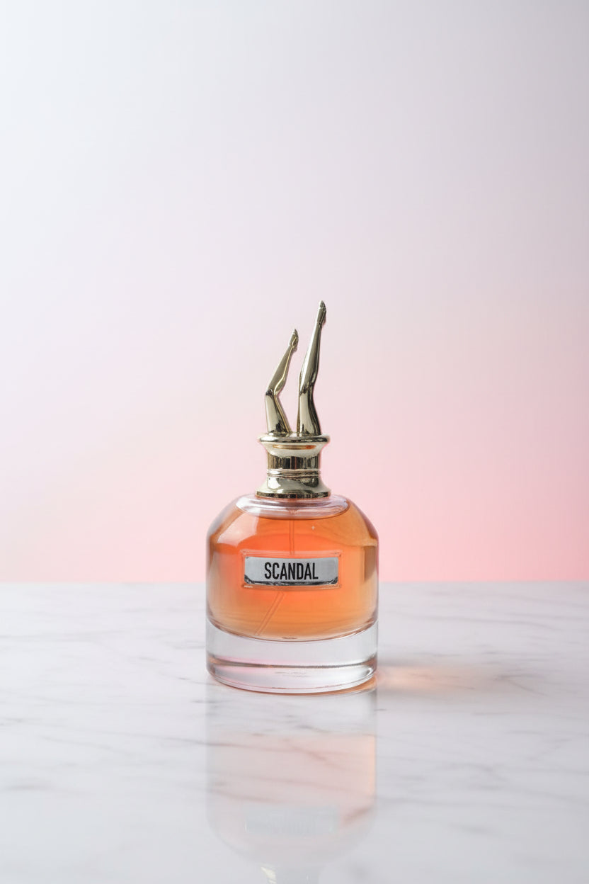 A hand holds a bottle of Scandal Kinfolk perfume with amber liquid and a gold spiky cap, with a pink box and a large woven circular decor piece in the background.