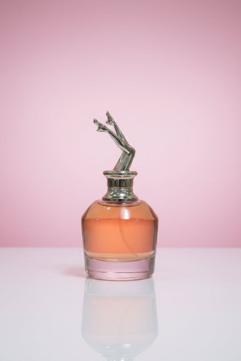 A hand holds a glass perfume bottle filled with pink liquid, featuring a metallic gold figure-shaped cap. A pink box labeled 'SCANDAL' is visible in the foreground, with a large woven gold circular decor piece behind the product.