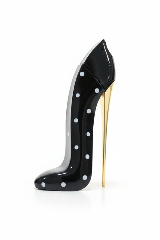 A hand with black nail polish holds a glossy black perfume bottle shaped like a stiletto heel with white polka dots and a gold sole, next to a boxed fragrance labeled Good Girl by Chiara Mumentara.