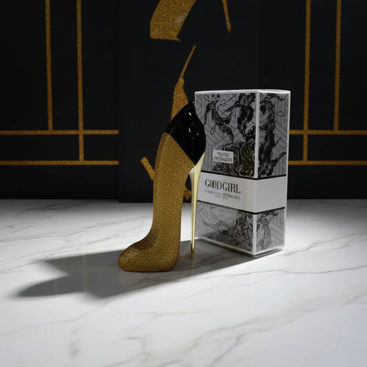 A gold glitter high-heeled shoe-shaped perfume bottle next to its illustrated product box labeled 'GOOD GIRL' and 'TESTER PRÉMISOR' on a reflective glass table with gold accents.