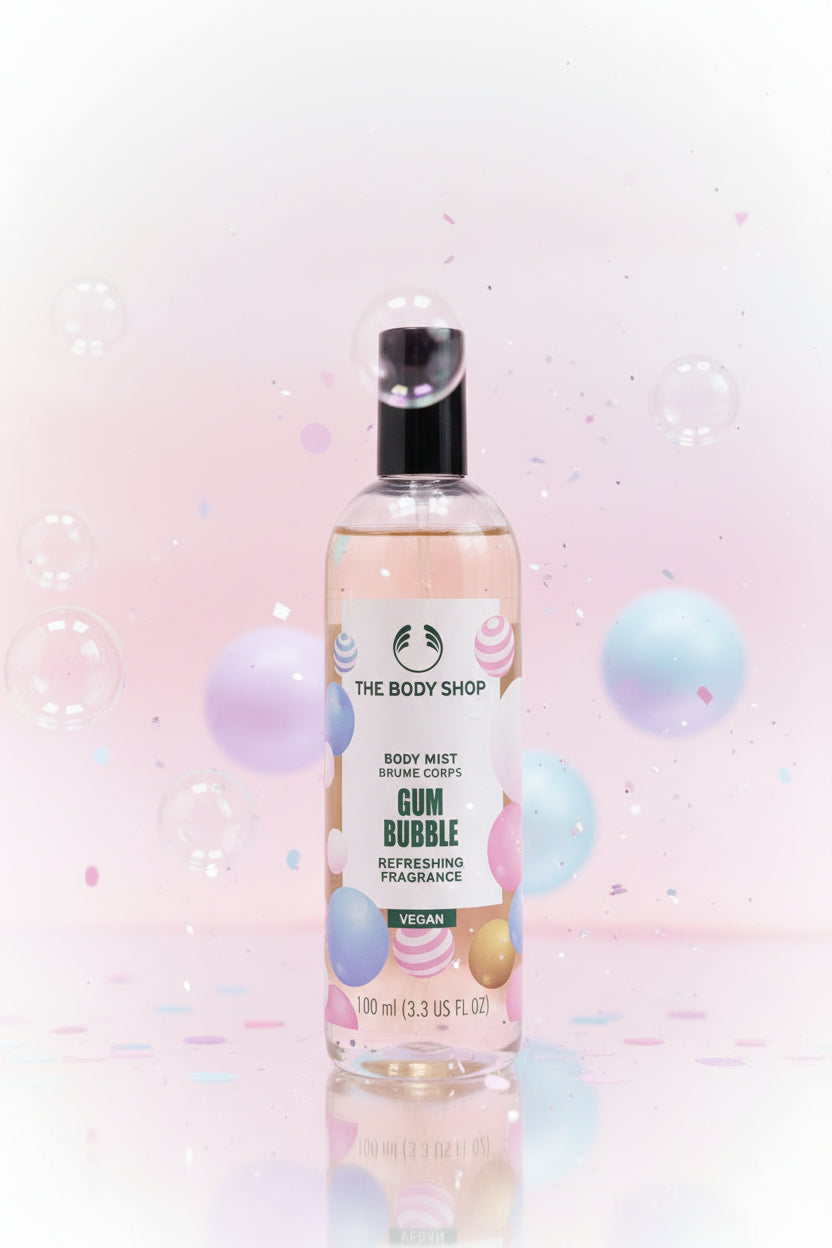 A bottle of The Body Shop Gum Bubble body mist fragrance, filled with a pale yellow liquid and featuring a black cap, set on a wooden table with a blurred home background.